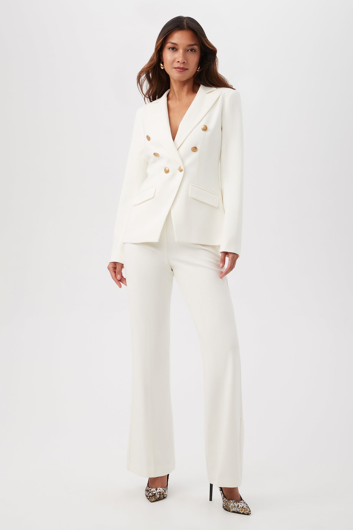 A woman stands against a plain background wearing the Midlands Luxe Drape Stretch Blazer Jacket in white, featuring gold buttons and wide-leg trousers, paired with leopard print high heels.