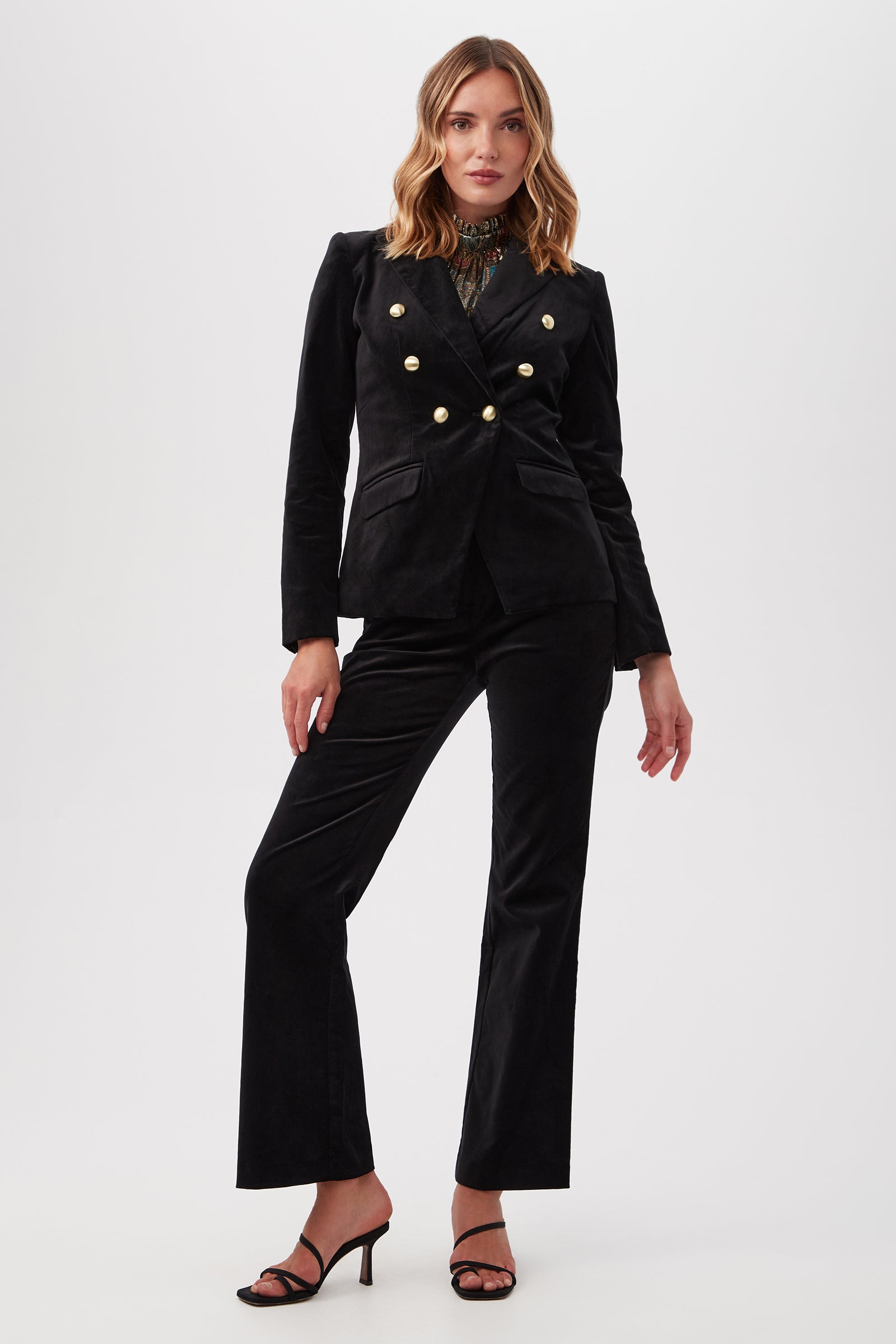 A woman stands confidently in the Midlands Velveteen Blazer Jacket with gold buttons, flared pants, and black heeled sandals against a plain white background.