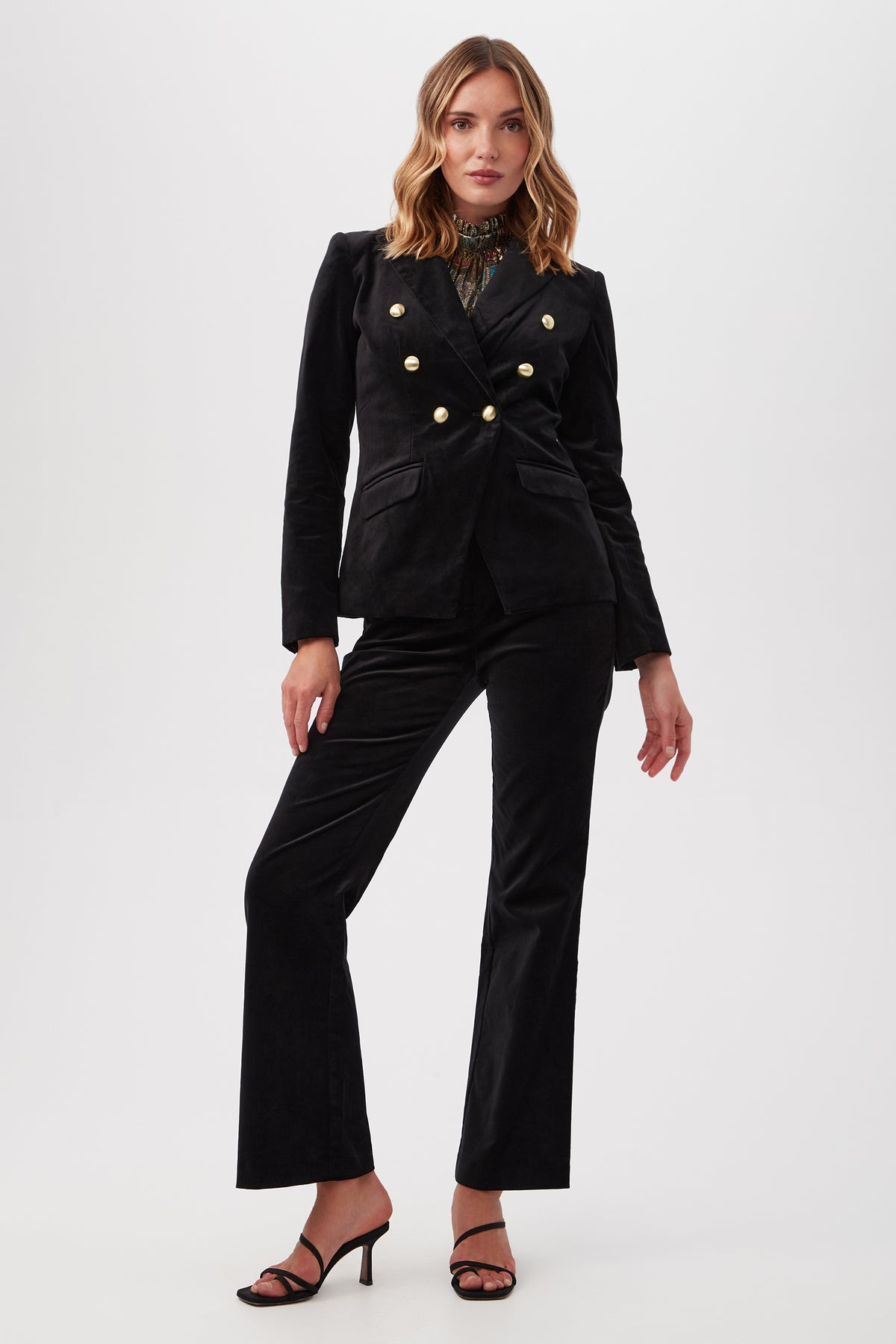 A woman stands confidently in Westport Velveteen Bootcut Pants, paired with a black double-breasted velvet blazer with gold buttons and black high-heeled sandals, set against a plain white background.