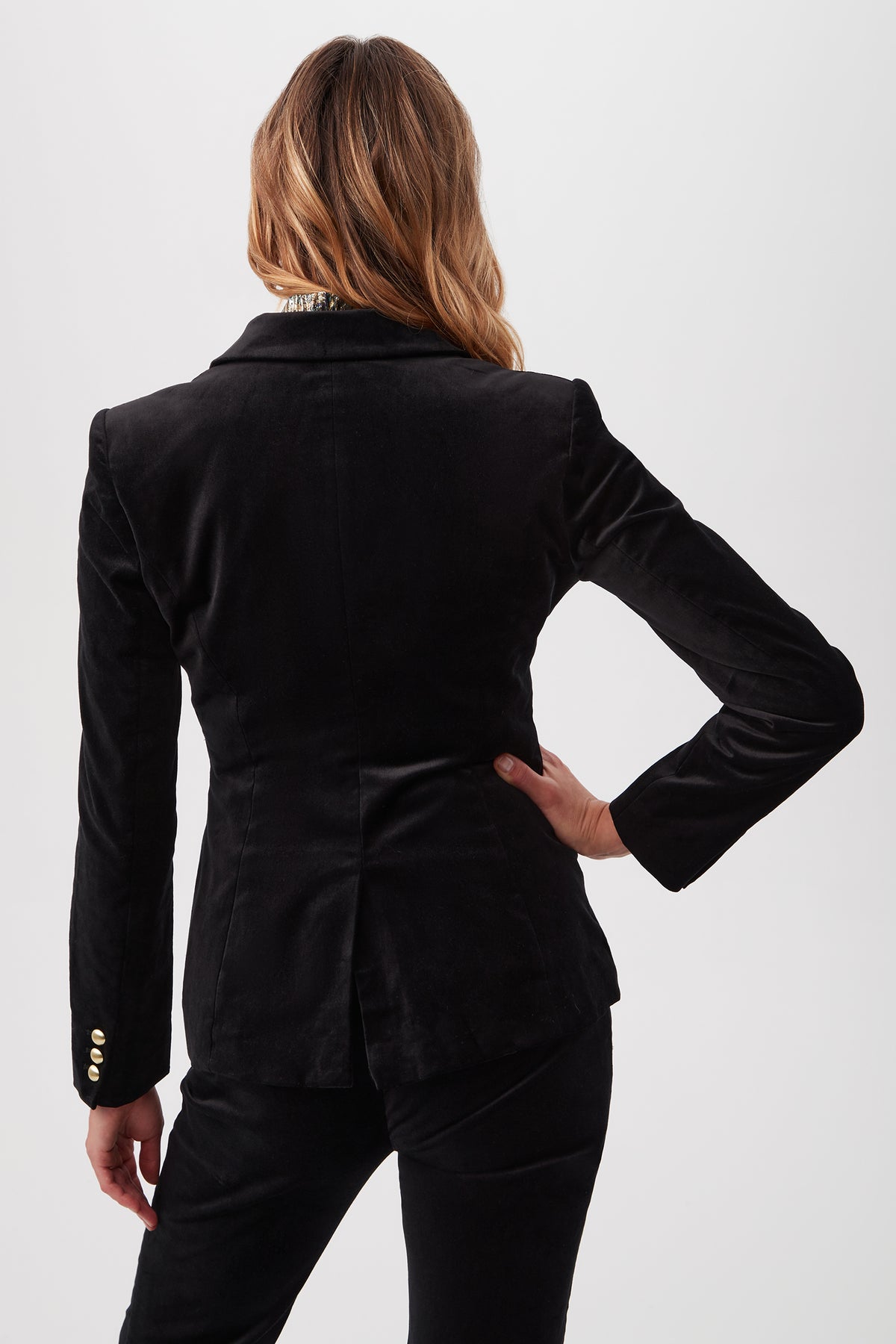 A person with wavy, light brown hair is seen from behind wearing the Midlands Velveteen Blazer Jacket in black, featuring gold sleeve buttons and matching pants. One hand rests on their hip against a plain white background.