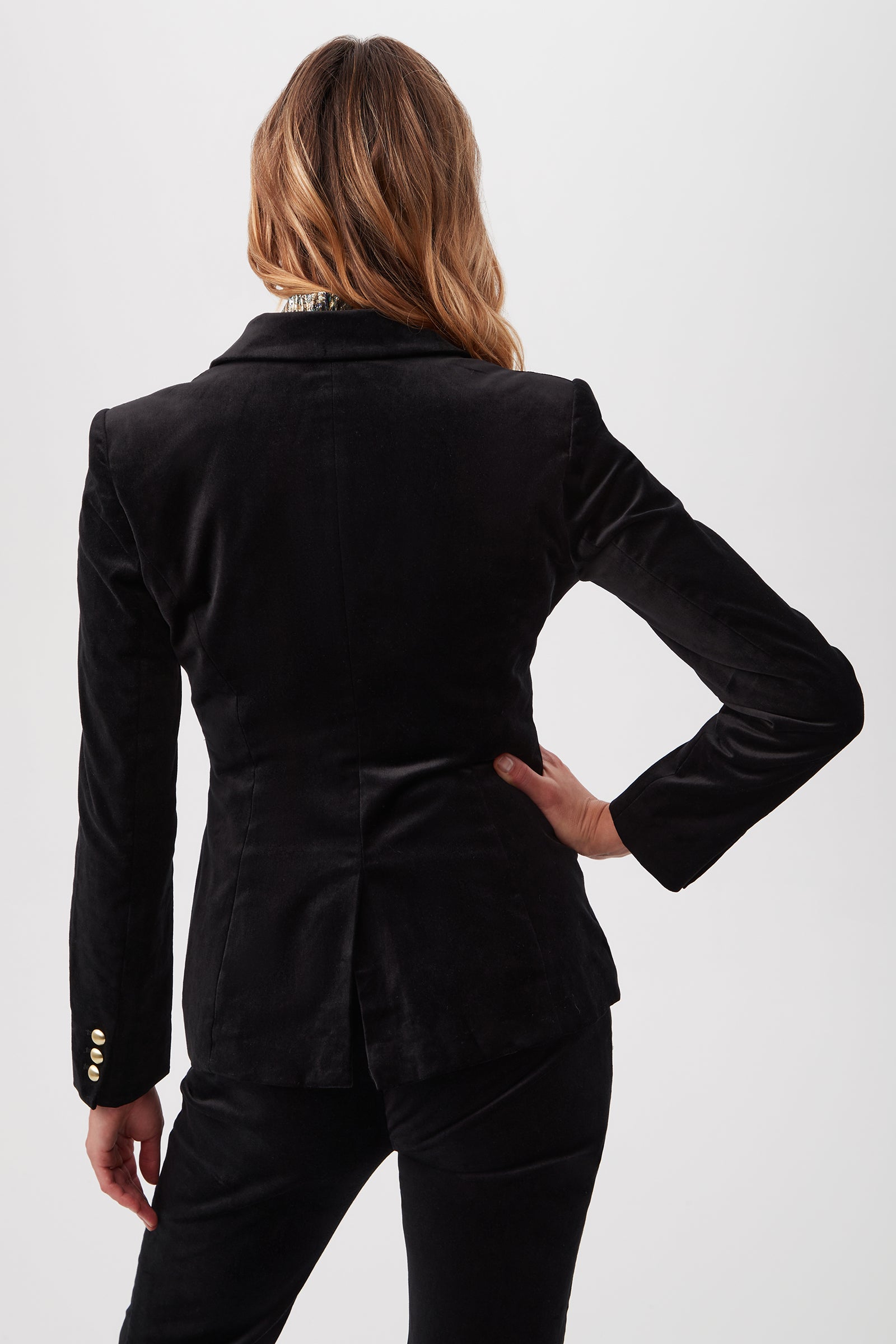 A person with wavy, light brown hair is seen from behind wearing the Midlands Velveteen Blazer Jacket in black, featuring gold sleeve buttons and matching pants. One hand rests on their hip against a plain white background.