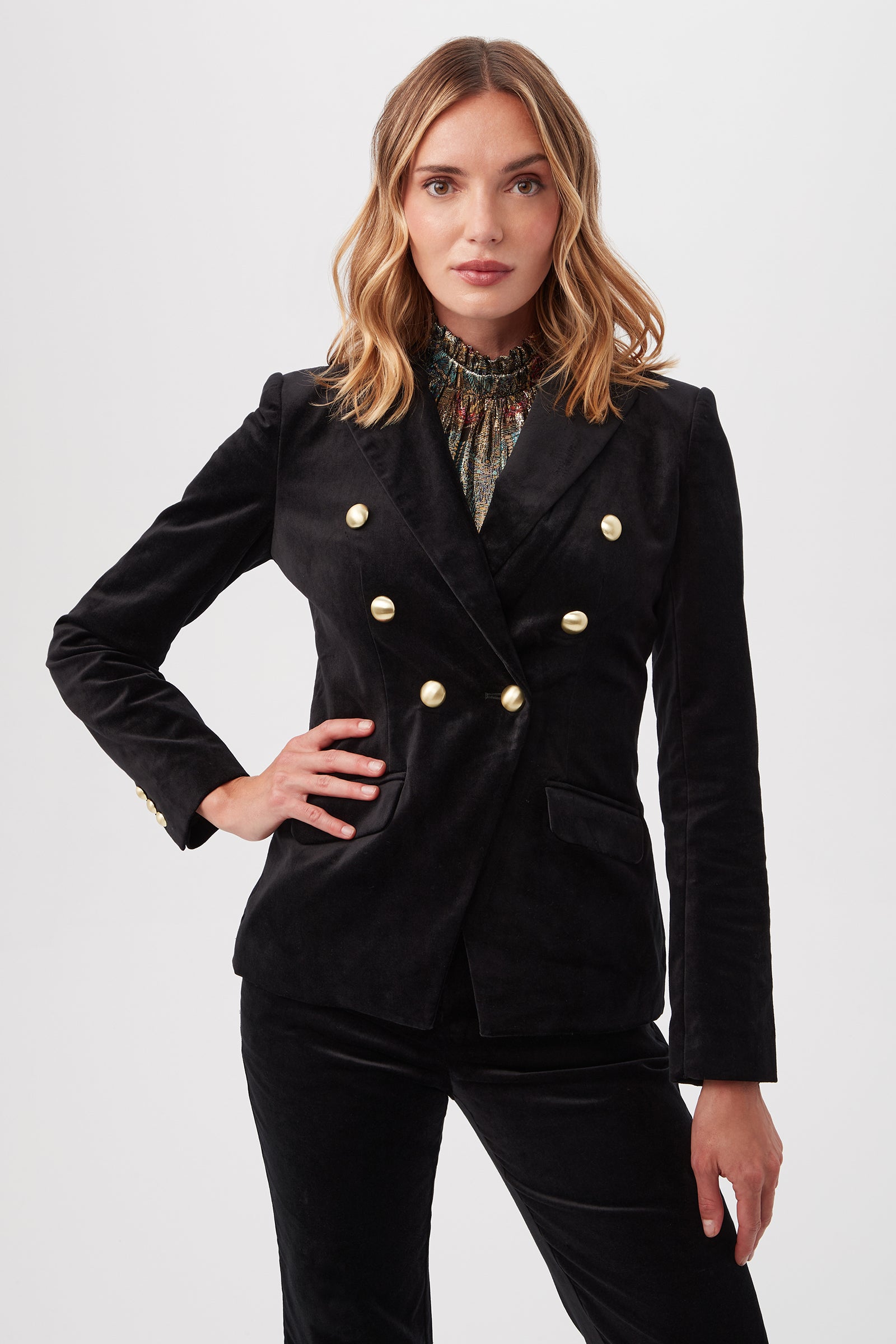 A woman poses with one hand on her hip against a plain white background, wearing the Midlands Velveteen Blazer Jacket in black with gold buttons and matching pants.