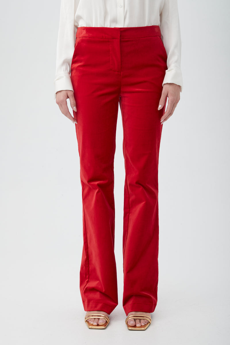 A person wearing the Westport Velveteen Bootcut Pant in bright red, paired with a white long-sleeve shirt and beige open-toe heeled sandals, stands against a plain white background with only the lower half of the body visible.