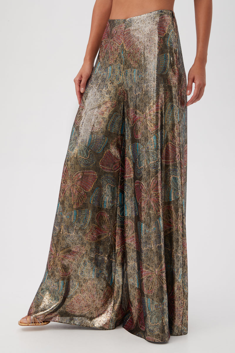 A person wearing the Siya Butterfly Print Silk Wide Leg Pant, featuring a metallic sheen with multicolored floral and abstract patterns, stands against a plain light background.