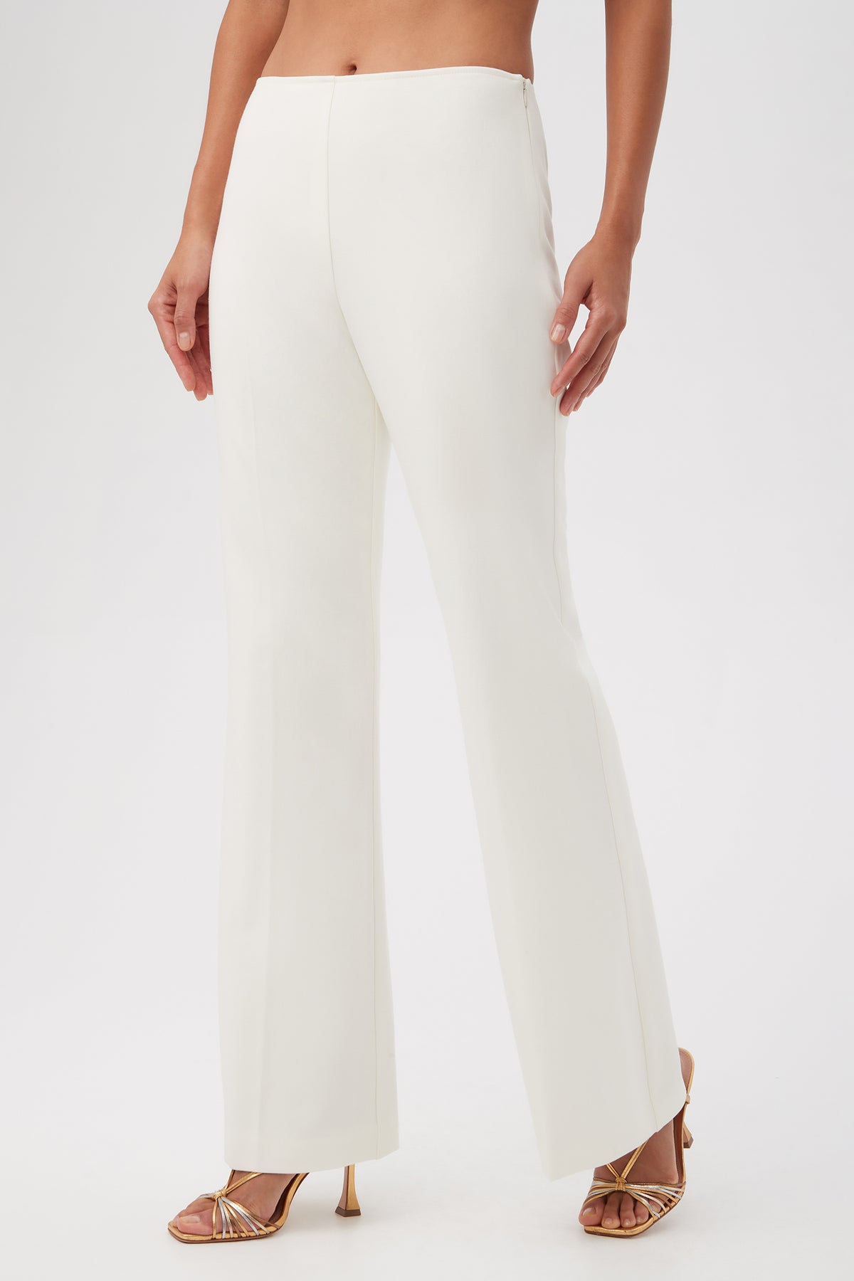 A person wears the Lancelot 2 Luxe Drape Bootcut Pants in high-waisted white, paired with gold strappy heels, standing against a plain white background. Photo is cropped at the chest.