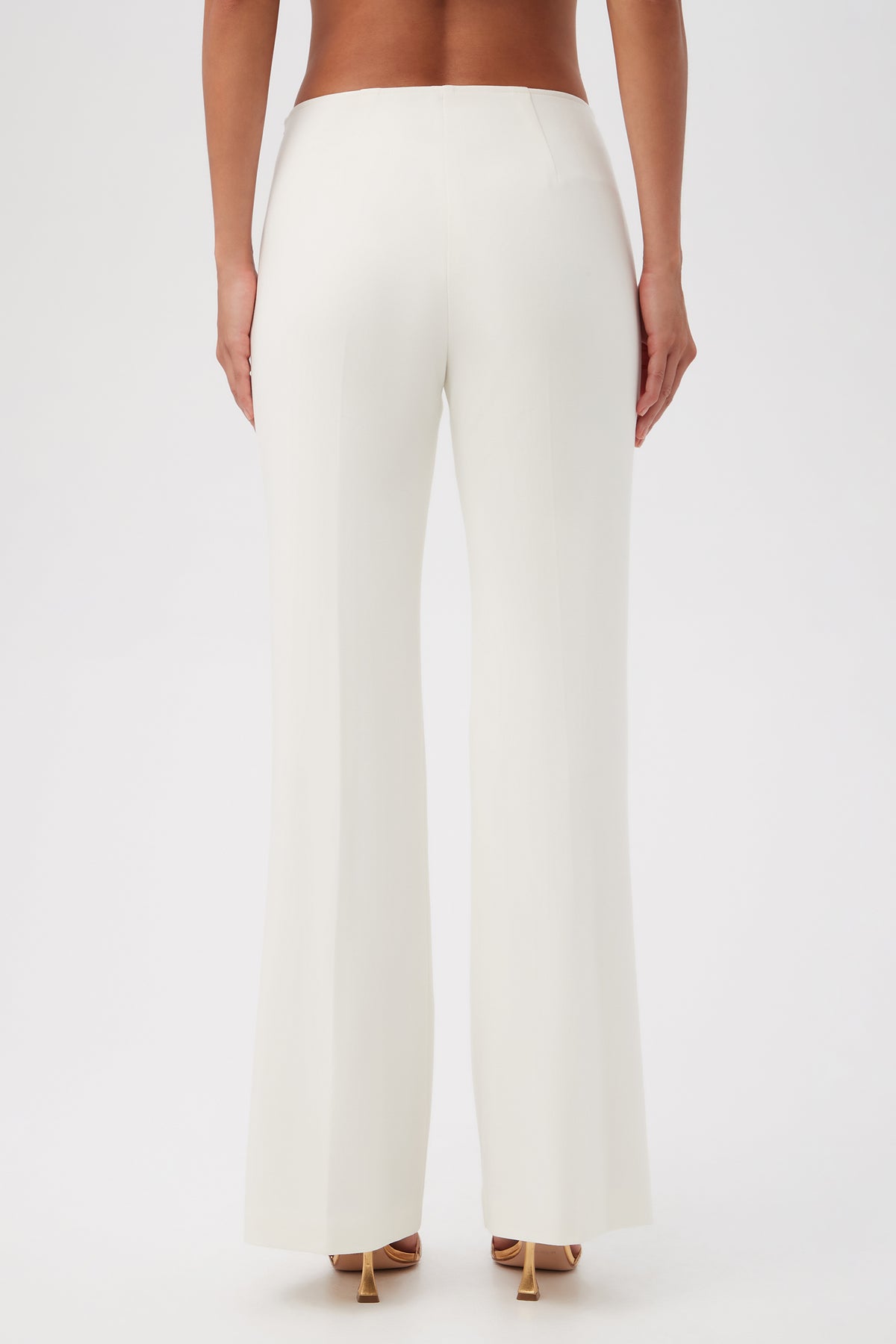 A person is shown from the back, wearing Lancelot 2 Luxe Drape Bootcut Pants in white with nude high-heeled sandals against a plain white background.