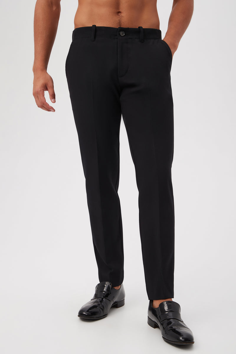 A man wearing the Alex Slim Trouser - Luxe Drape in BLACK, with his hands in the pockets and black shiny dress shoes, stands against a plain light background.