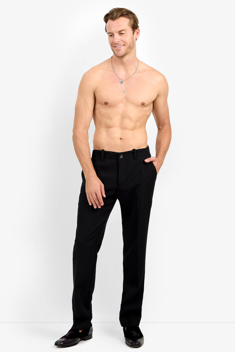 A shirtless man wears the Alex Slim Trouser - Luxe Drape in black with matching shoes, standing against a plain white backdrop, one hand in his pocket and smiling slightly.