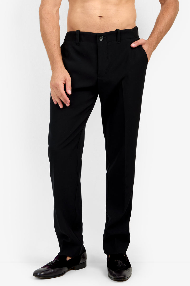 A man wears the Alex Slim Trouser - Luxe Drape in black with black shoes, standing against a white background, shirtless with one hand in his pocket.