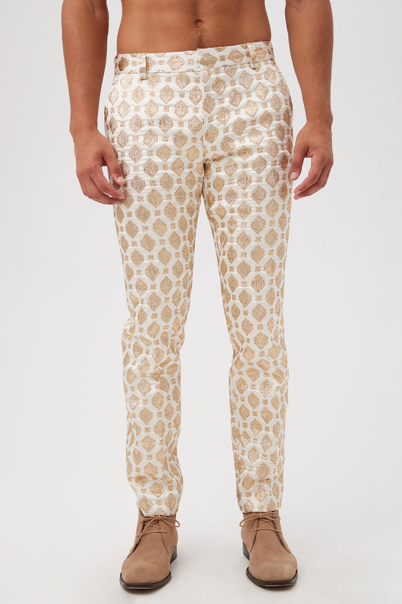 A person wears the Clyde Slim Trouser - Jubilant Jacquard, featuring shiny white fabric with a gold geometric pattern and beige dress shoes, standing against a plain white background with a bare torso.