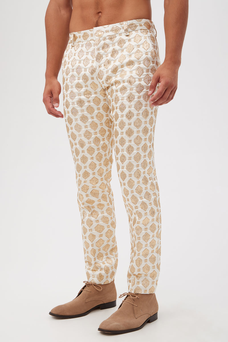 A person wears the Clyde Slim Trouser - Jubilant Jacquard, featuring a gold geometric pattern on white, paired with light brown suede lace-up shoes and standing against a plain white background. The upper body is not visible.