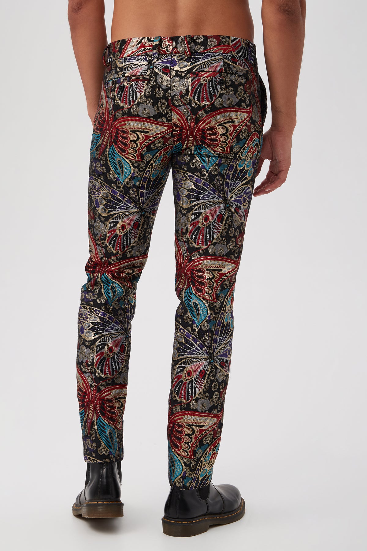 A man, shirtless and in black shoes, stands with his back to the camera wearing the Clyde Slim Trouser in Monarch Jacquardâ€”a vibrant, multicolored butterfly and floral patterned slim-fit trouserâ€”set against a plain white background.