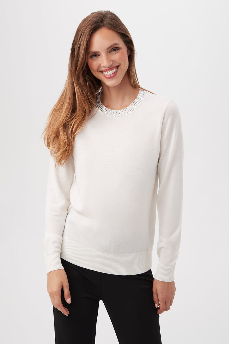 A woman with long brown hair is smiling and wearing the Laelia Merino Wool Sweater With Rhinestone Trim in WINTER WHITE, paired with black pants. She stands against a plain white background.