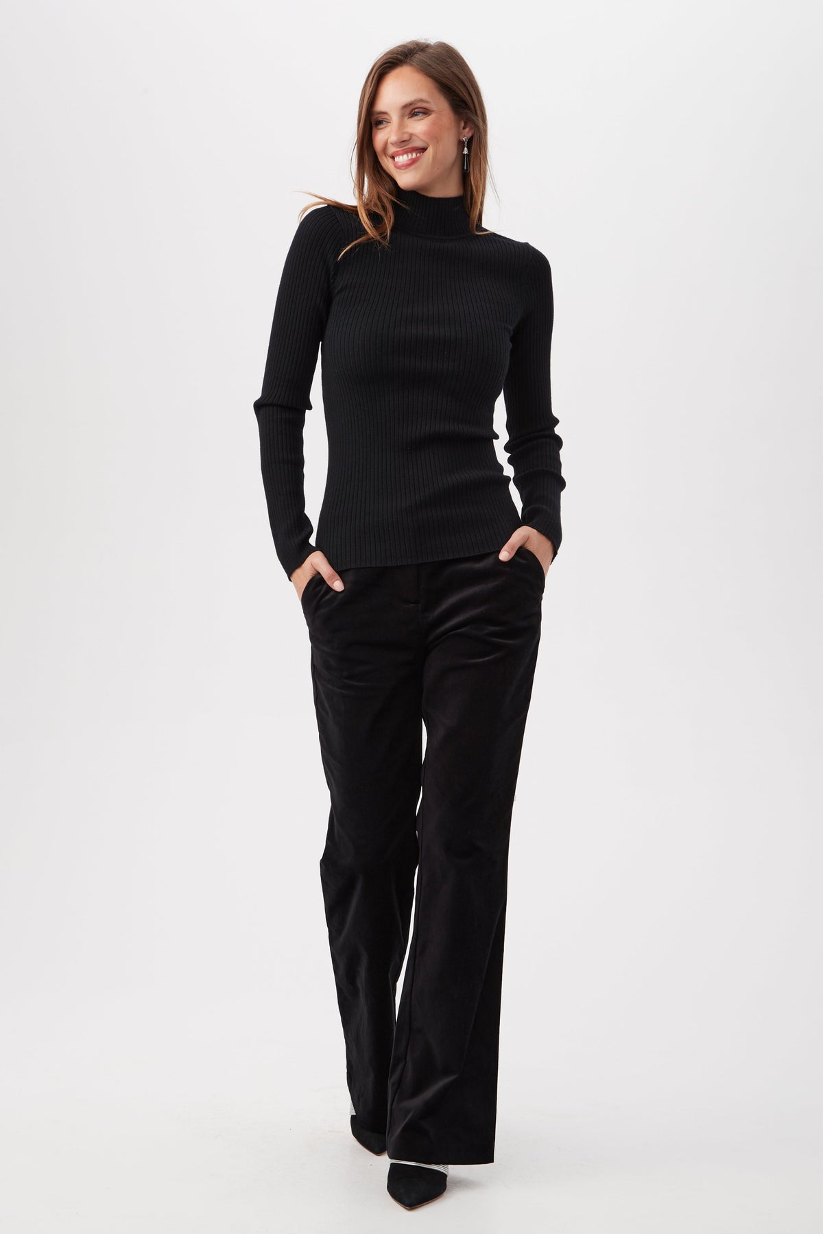 A woman smiles with her hands in her pockets, wearing the Sade Merino Wool Mockneck Sweater With Open Back and loose black pants against a plain white background.