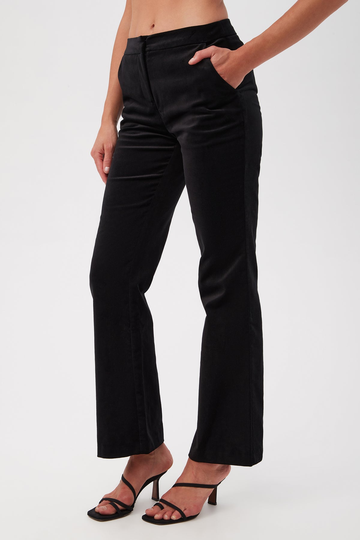 A person stands against a plain background wearing Westport Velveteen Bootcut Pants and black strappy heeled sandals, with one hand in their pocket and their midriff visible.
