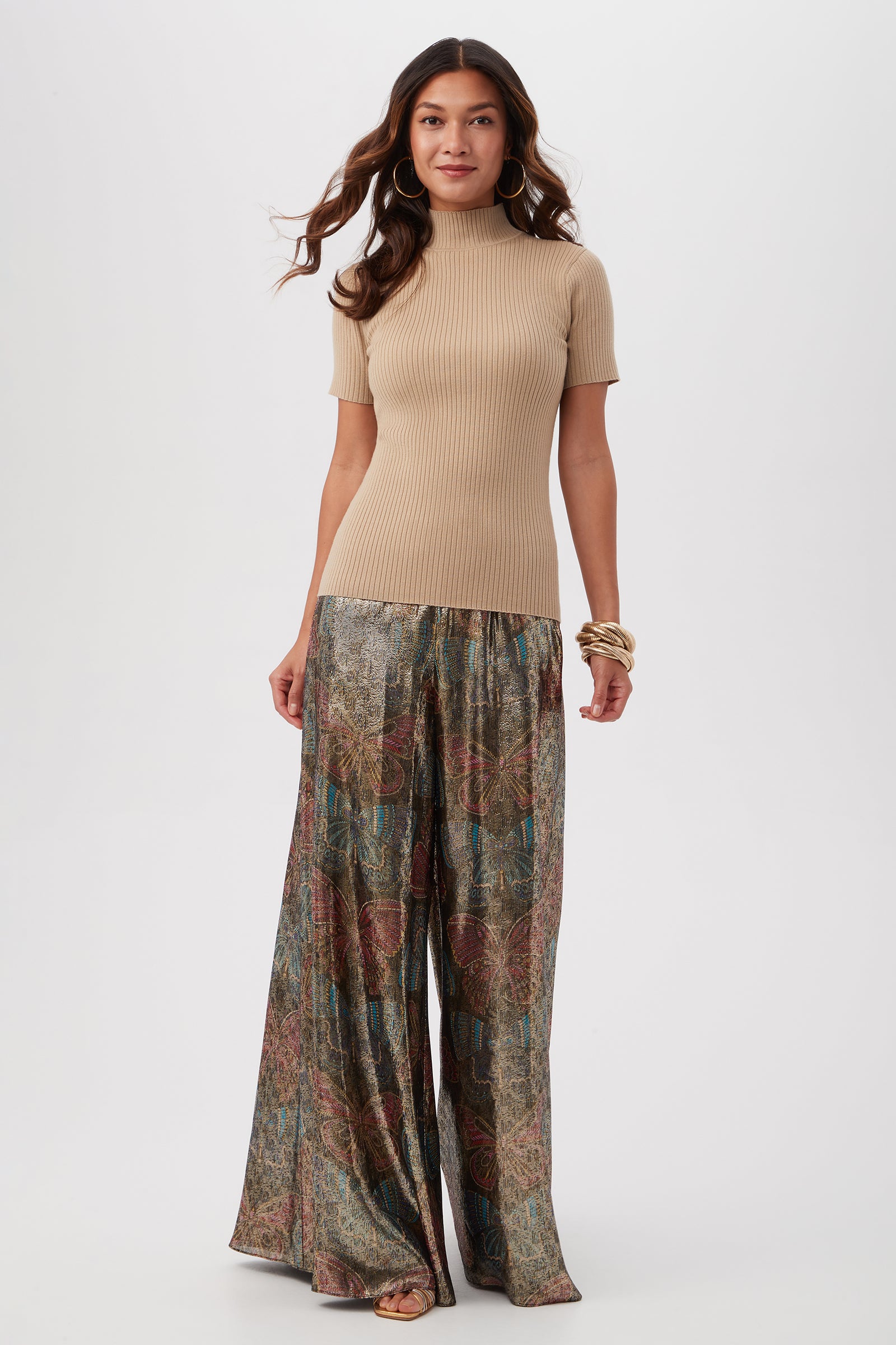 A woman with long wavy hair wears the Armeni Merino Wool Short Sleeve Mockneck Top in beige and wide-leg, patterned metallic pants in earthy tones, accessorized with hoop earrings and bangles, against a plain background.