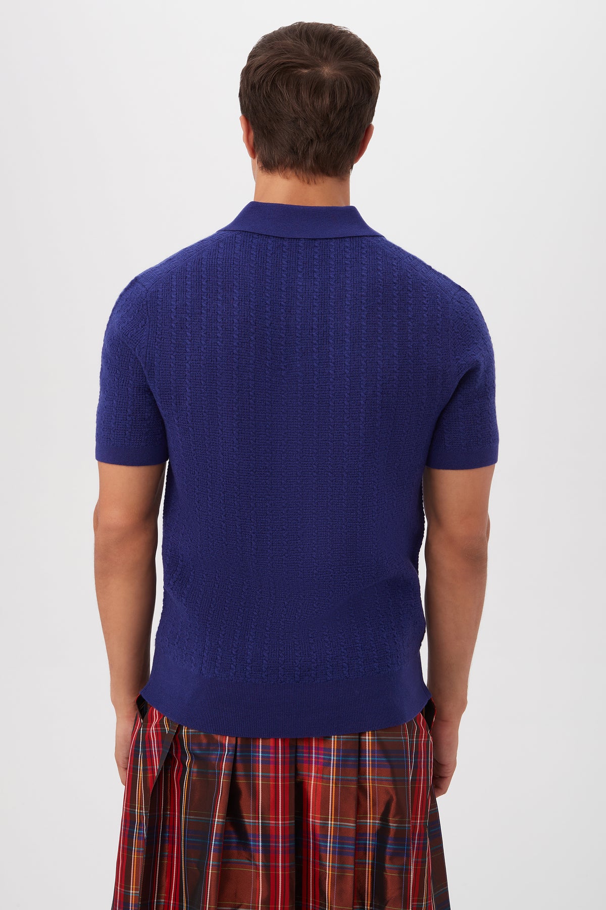 A person is seen from behind wearing the Asher Short-Sleeve Polo - Merino Wool in cobalt blue with a red plaid skirt against a plain white background.
