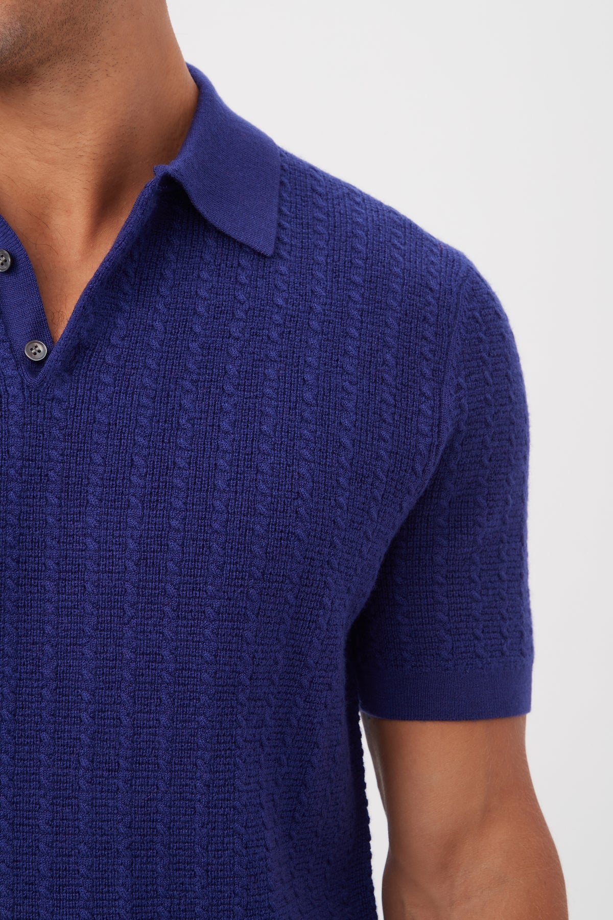 A person wears the Asher Short-Sleeve Polo - Merino Wool, a textured deep blue knit polo with a collar and buttons, shown from shoulders to waist against a plain white background.