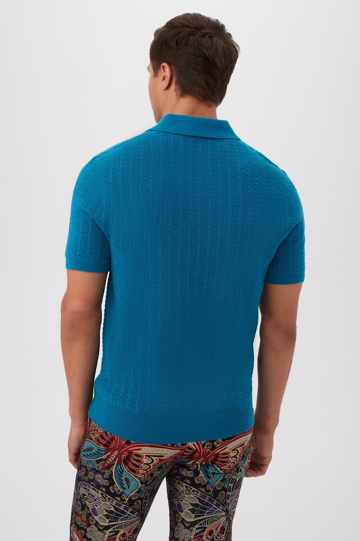 A man faces away, wearing the Asher Short-Sleeve Polo in textured blue merino wool and colorful paisley-patterned pants, set against a plain white background.