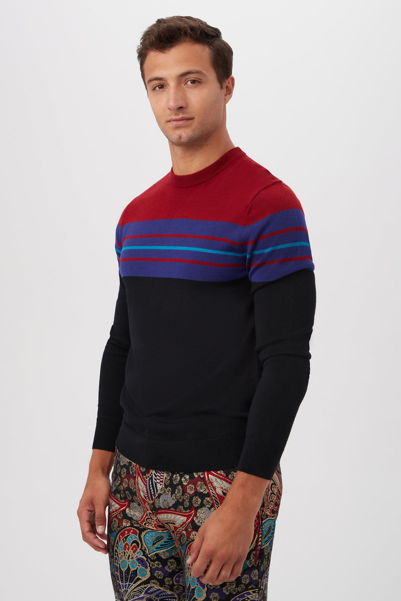 A man poses against a white background wearing the Romero Crewneck Sweater, a black knit with red and blue horizontal stripes on the chest and shoulders, paired with patterned pants featuring colorful, intricate designs.