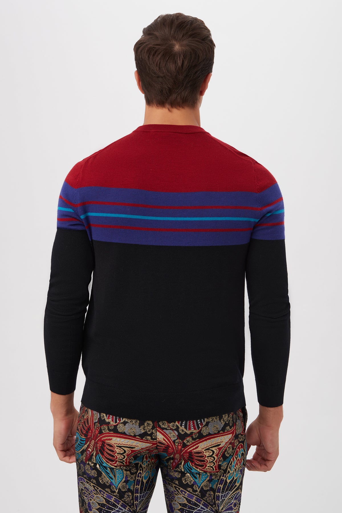 A man stands with his back to the camera, wearing the Romero Crewneck Sweaterâ€”featuring red and black sections with horizontal blue and purple stripesâ€”paired with butterfly-print pants.