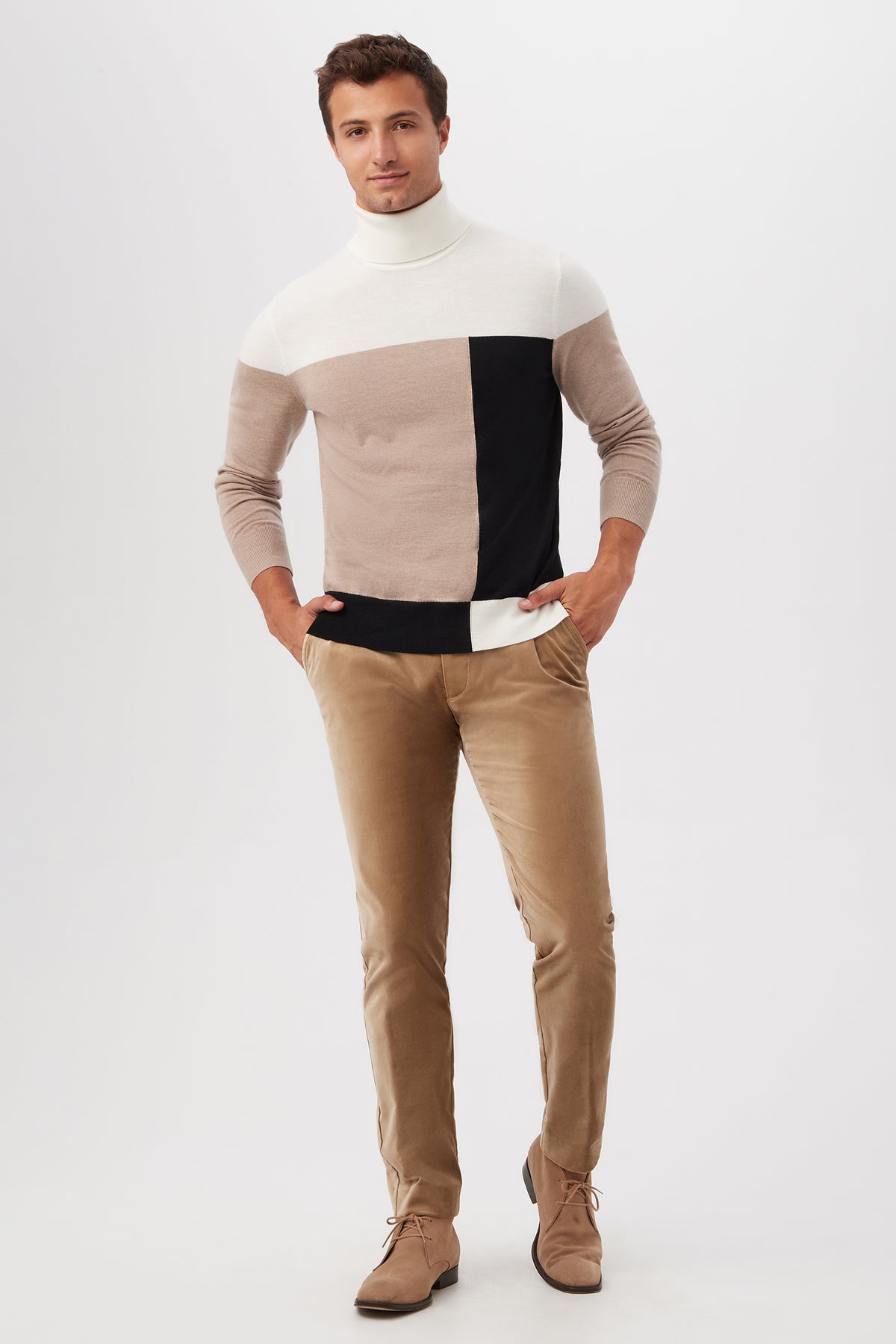 A man with short brown hair and a relaxed expression wears the Callisto Turtleneck Sweater in merino wool, featuring white, beige, and black color-block sections, paired with tan pants and light brown shoes against a white background.