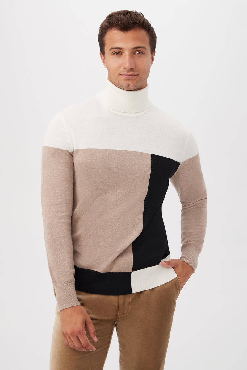 A man poses against a plain white background wearing the Callisto Turtleneck Sweater in Multi, featuring white, beige, and black color-block sections, with one hand in his pocket.