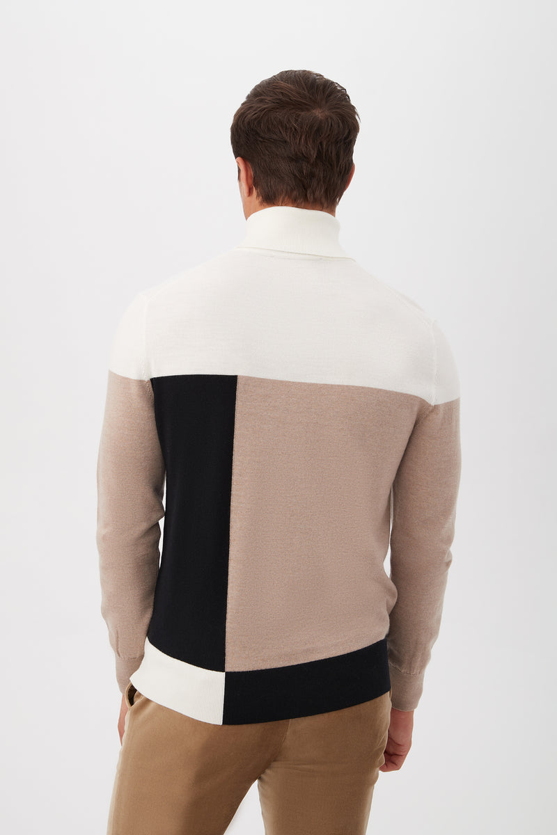 A man stands with his back to the camera, wearing the Callisto Turtleneck Sweater in Merino Woolâ€”white, black, and beige color-blockedâ€”paired with light brown pants against a plain white background.
