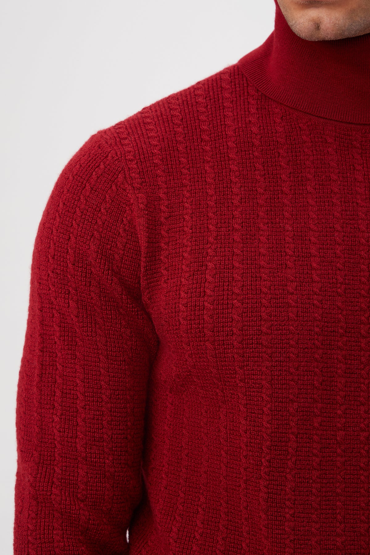 Close-up of a person wearing the Woodrow Turtleneck Sweater â€“ Merino Wool in red, highlighting its textured cable-knit pattern and fabric detail. The persons head is partially out of frame.