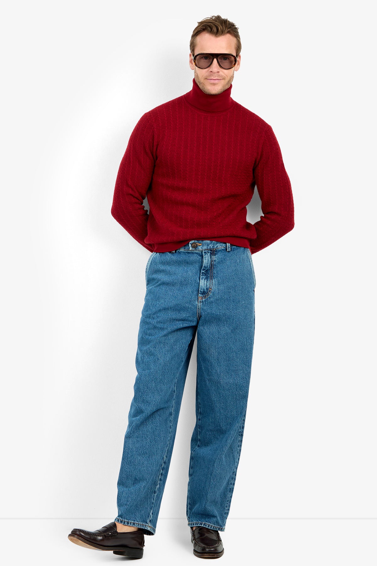 A man wearing the Woodrow Turtleneck Sweater - Merino Wool in red, blue jeans, brown loafers, and sunglasses stands against a white background with his hands behind his back.