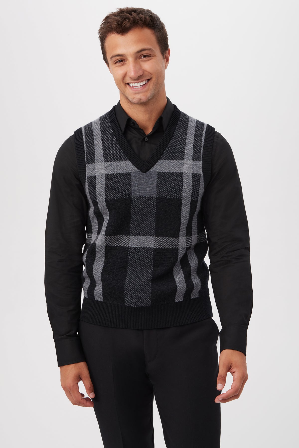 A smiling man stands against a plain white background, wearing a black dress shirt and pants with the Neso Plaid Knit Sweater Vest made of Merino Wool.