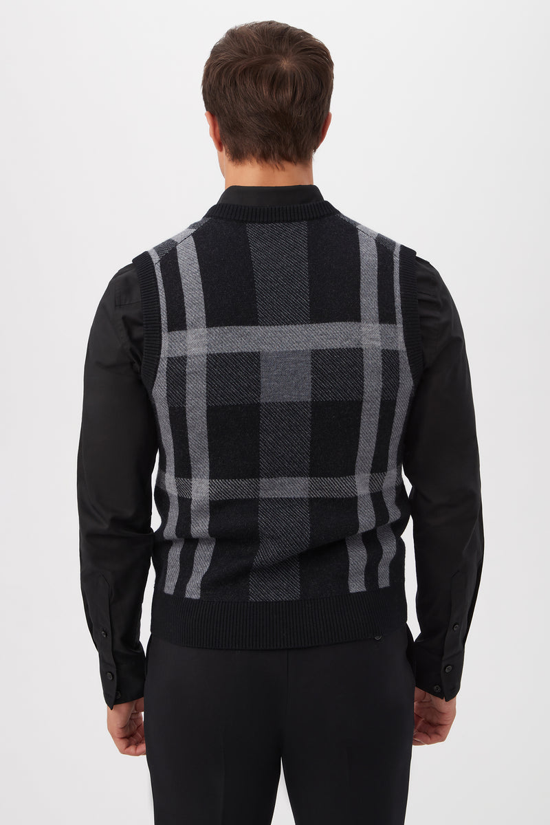 A man with short brown hair is seen from behind, wearing a black long-sleeve shirt and the Neso Plaid Knit Sweater Vest - Merino Wool in black and gray, standing against a plain white background.