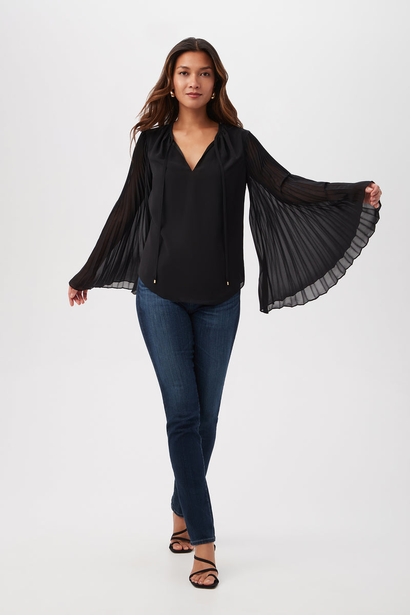A woman poses against a white background wearing the Golden Root Split V-Neck Top with accordion-pleated long sleeves, paired with blue jeans and black heeled sandals, walking forward with arms extended to highlight the distinctive sleeves.