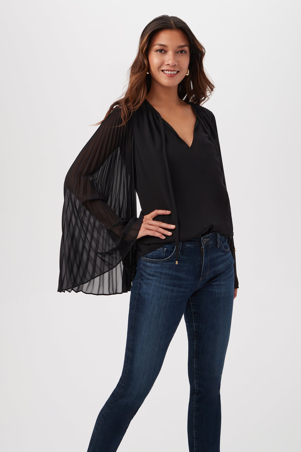 A woman smiles in the Golden Root Split V-Neck Top With Accordion-Pleated Long Sleeves in black, paired with dark blue jeans. She has long, wavy hair and poses against a plain white background.