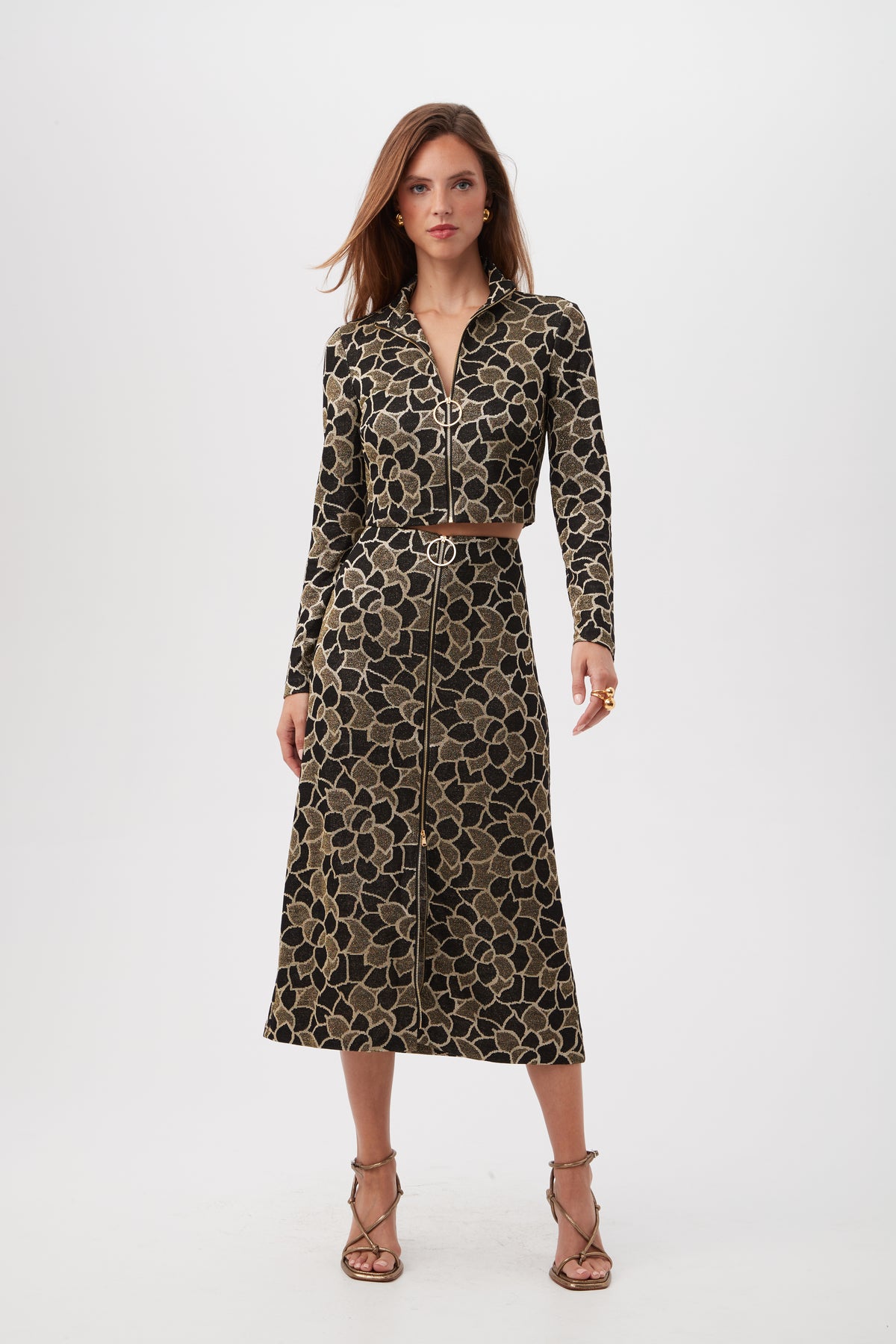 A woman stands against a white background wearing the Frosty Meadows Cropped Knit Jacquard Zip-Front Top with long sleeves and a floral pattern, her hair loose and paired with strappy high-heeled sandals.