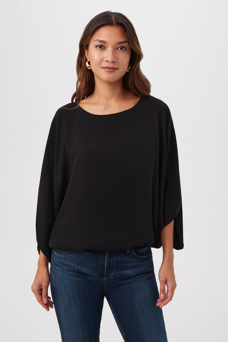 A woman with long brown hair, wearing the black Manhattan Relaxed Short Dolman Sleeve Top and blue jeans, stands facing the camera against a plain white background.