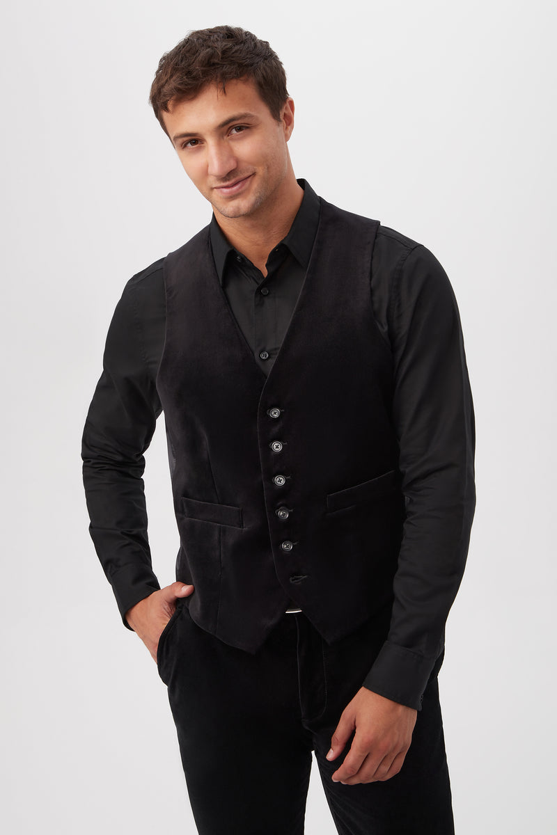 A man with short brown hair smiles with one hand in his pocket, wearing the Irving Vest - Black Velveteen over a black dress shirt and matching black pants against a plain light background.