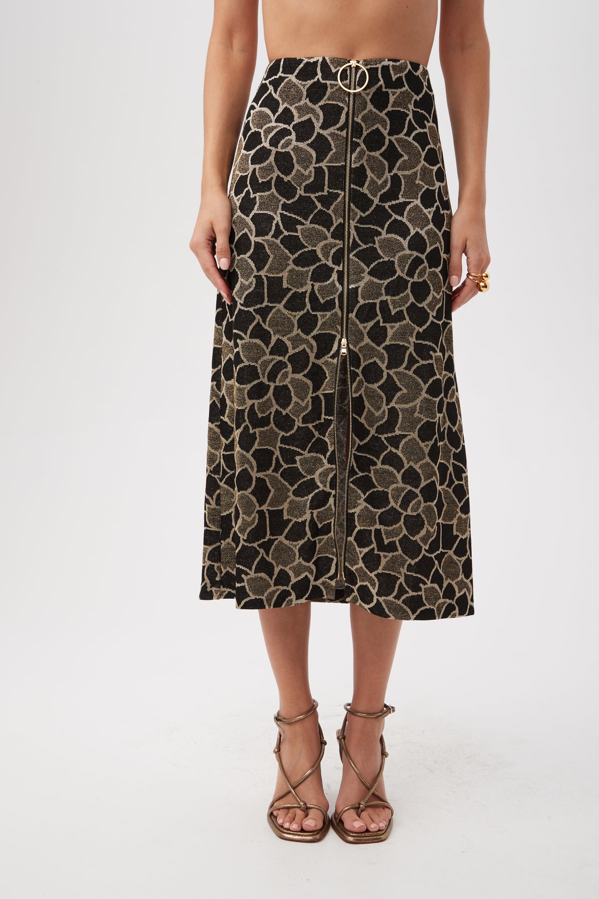 A woman wears the Zane Metallic Knit Jacquard Zip-Front Midi Skirt in multi, featuring a high waist, gold floral pattern, front zipper, and gold belt detail with strappy heeled sandals. Her upper body is not visible.