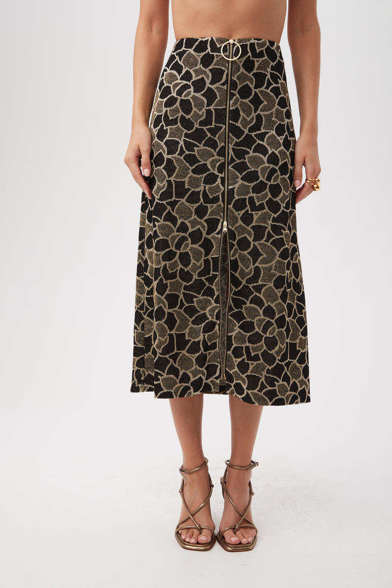 A woman wears the Zane Metallic Knit Jacquard Zip-Front Midi Skirt in multi, featuring a high waist, gold floral pattern, front zipper, and gold belt detail with strappy heeled sandals. Her upper body is not visible.