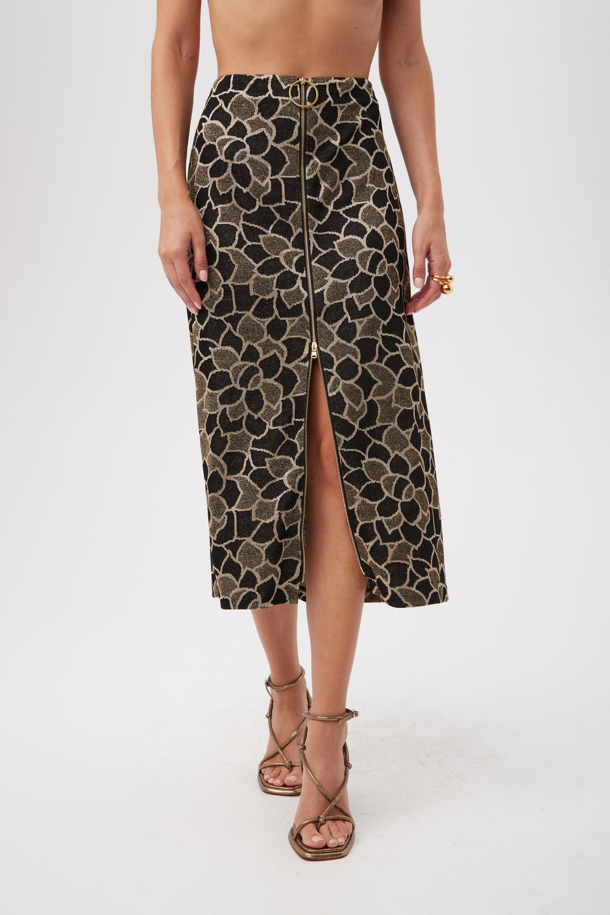 A person wears the Zane Metallic Knit Jacquard Zip-Front Midi Skirt in black and gold with a front slit, paired with strappy high-heeled sandals. The upper body is mostly out of frame.