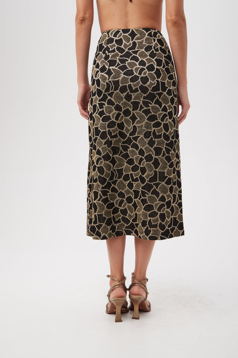 A woman faces away, wearing the Zane Metallic Knit Jacquard Zip-Front Midi Skirt in gold and black with strappy heeled sandals against a plain white background.