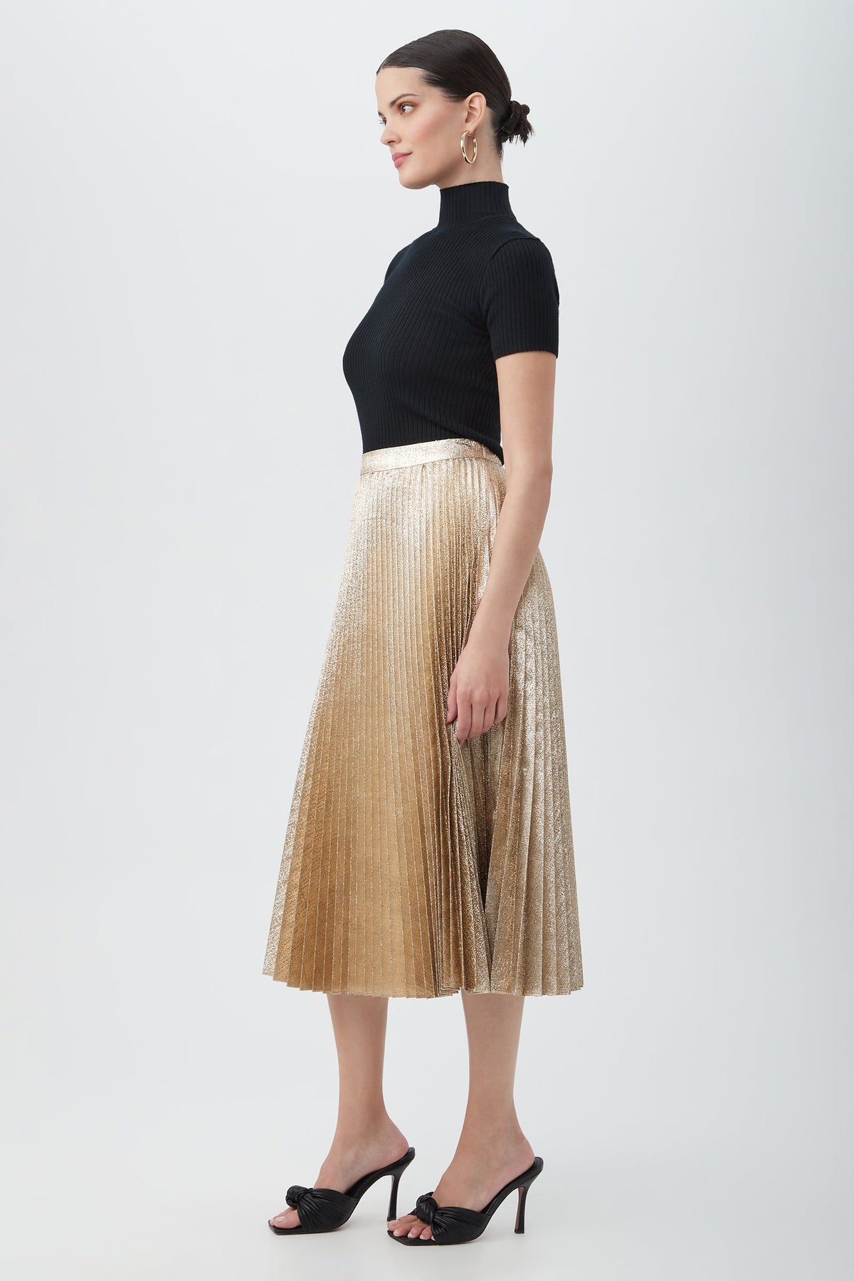 Woman in profile against a plain background, wearing a black short-sleeve turtleneck, the Faye Luminary LamâˆšÂ© Midi Skirt With Accordion Pleats in gold, black heeled sandals, gold hoop earrings, and her hair in a low bun.