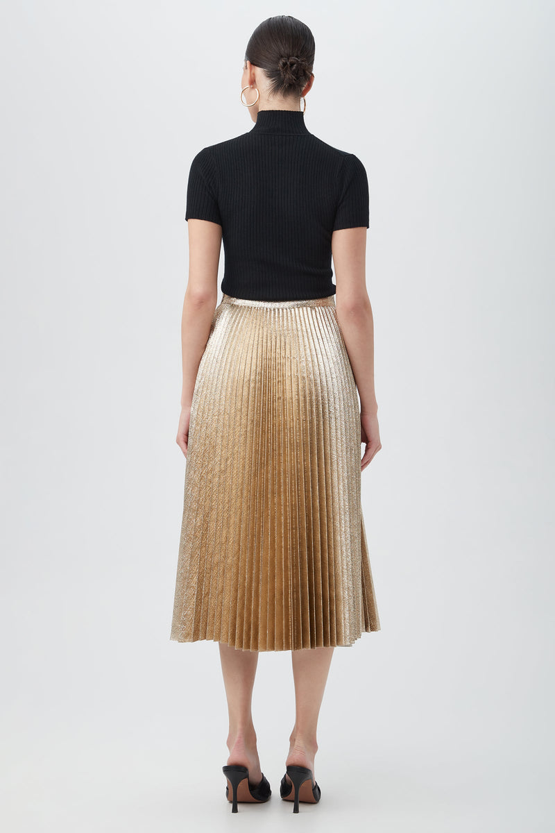 A woman with her hair in a sleek bun and large hoop earrings faces away, wearing a black ribbed short-sleeve top, black open-toe heels, and the Faye Luminary LamâˆšÂ© Midi Skirt With Accordion Pleats in metallic gold.