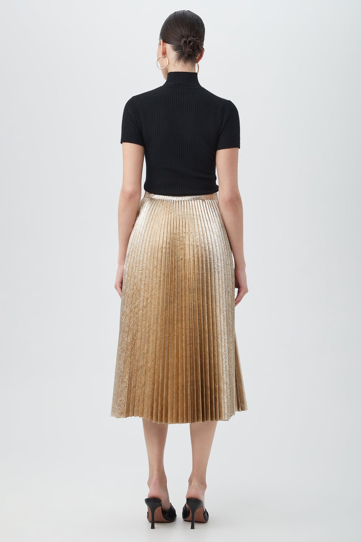 A woman with her hair in a sleek bun and large hoop earrings faces away, wearing a black ribbed short-sleeve top, black open-toe heels, and the Faye Luminary LamâˆšÂ© Midi Skirt With Accordion Pleats in metallic gold.