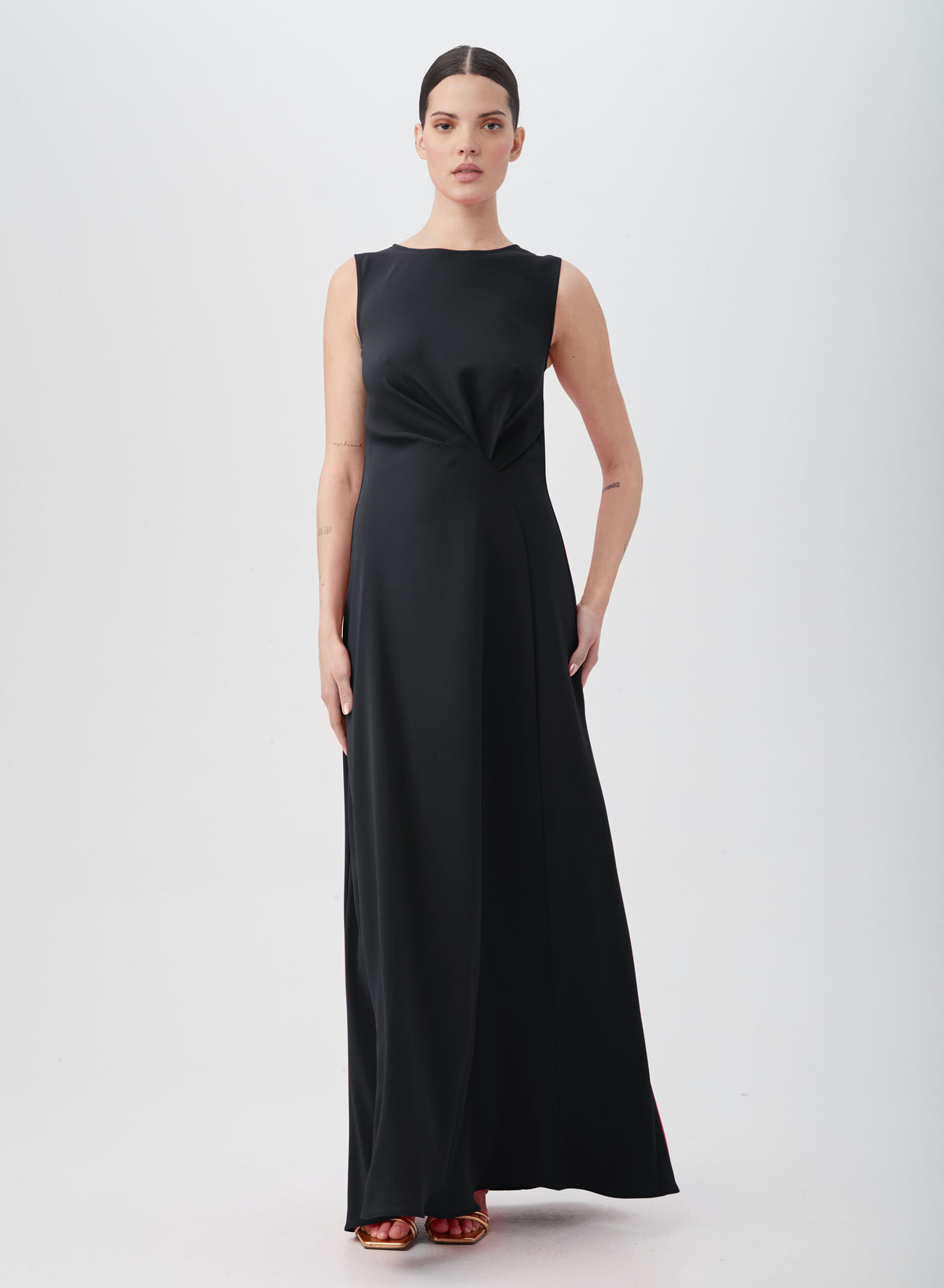 A woman stands against a plain background in the MYTHICAL DRESS, a sleeveless, floor-length black polyester gown with waist pleats. She wears red open-toe heels, has dark hair pulled back, and gazes directly at the camera.
