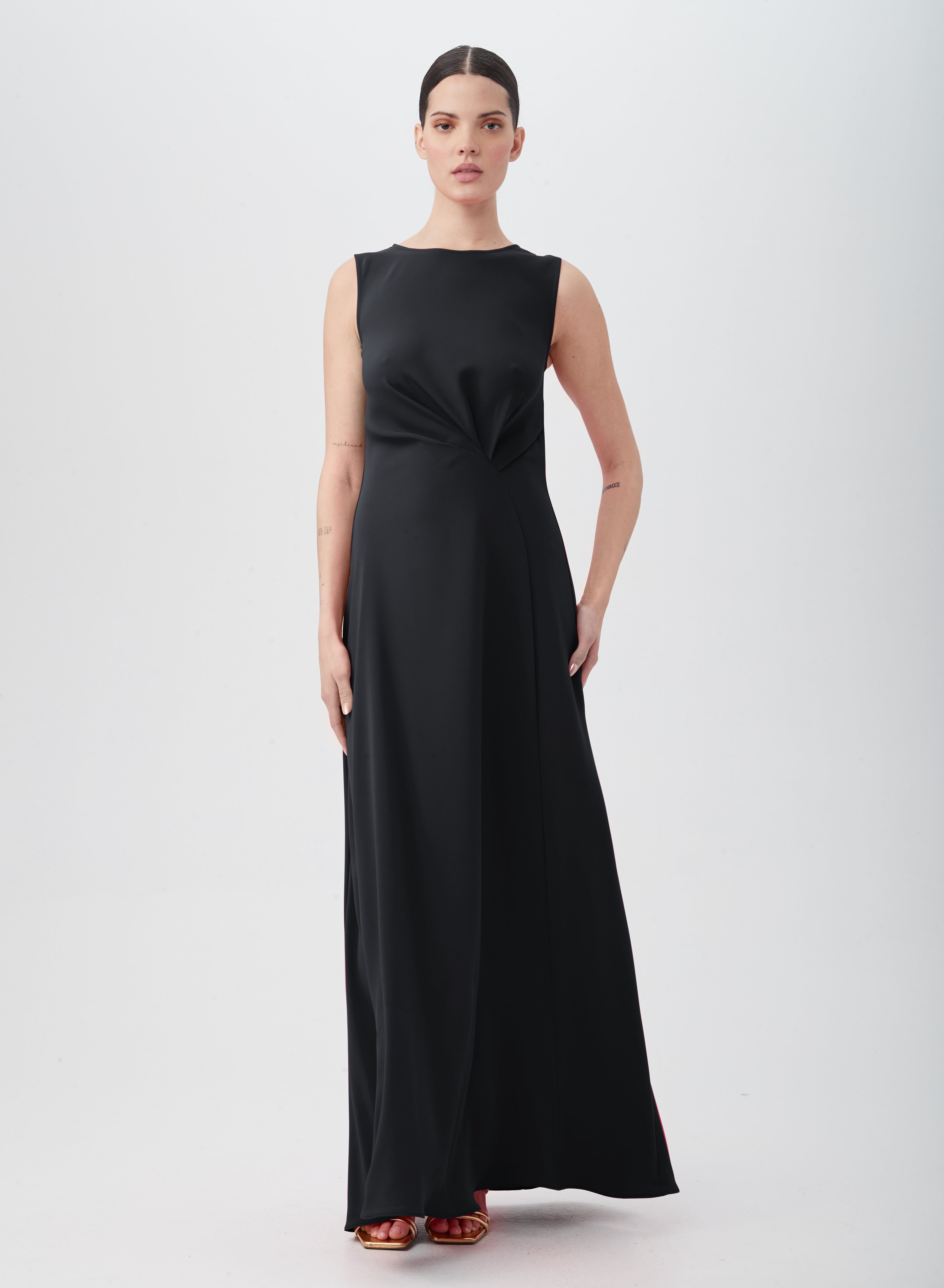 A woman stands against a plain background in the MYTHICAL DRESS, a sleeveless, floor-length black polyester gown with waist pleats. She wears red open-toe heels, has dark hair pulled back, and gazes directly at the camera.