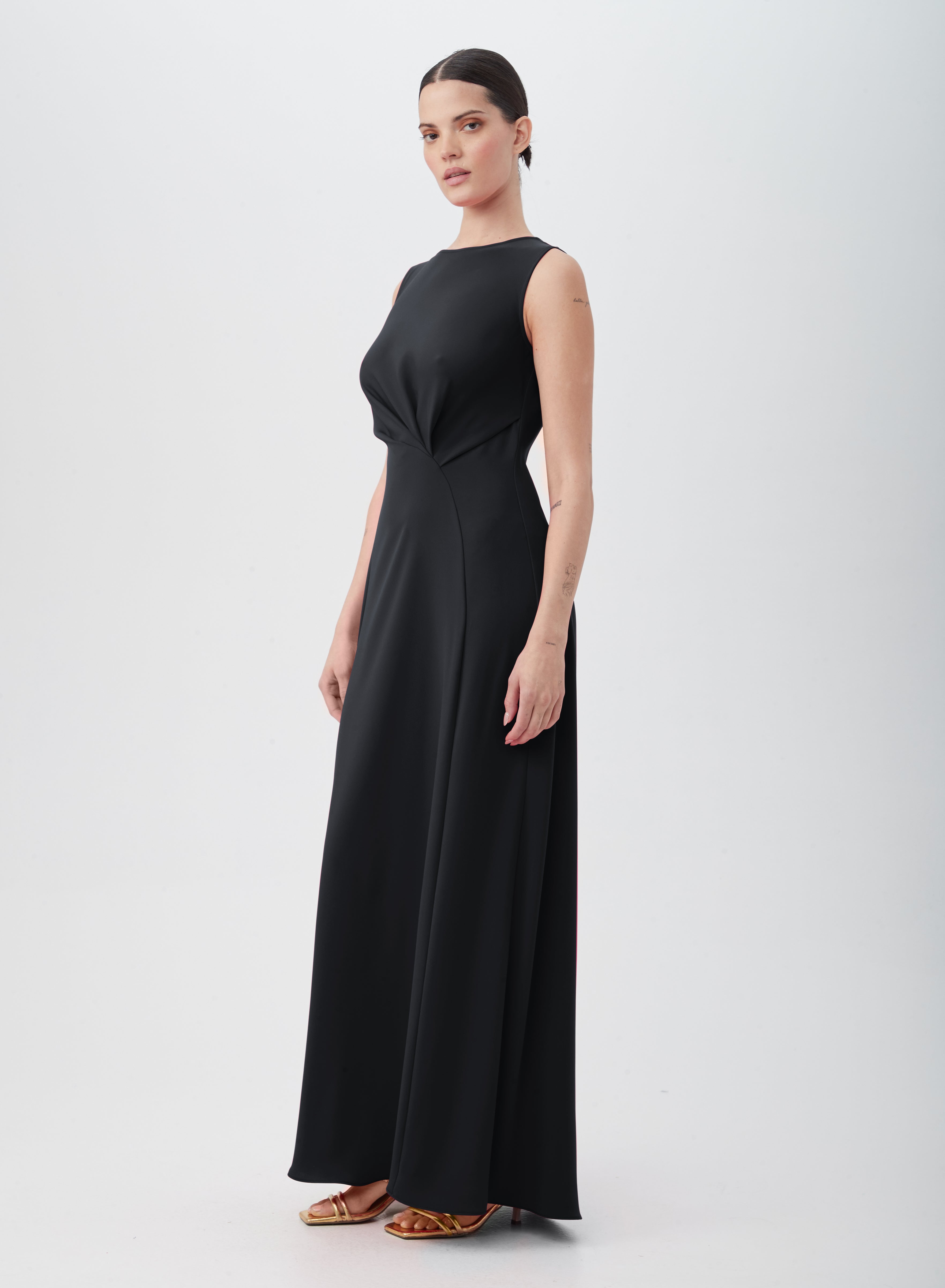 A woman wears the MYTHICAL DRESS, a sleeveless, floor-length black gown of 100% polyester with a fitted waist, paired with gold high-heeled sandals, posed against a plain white background.