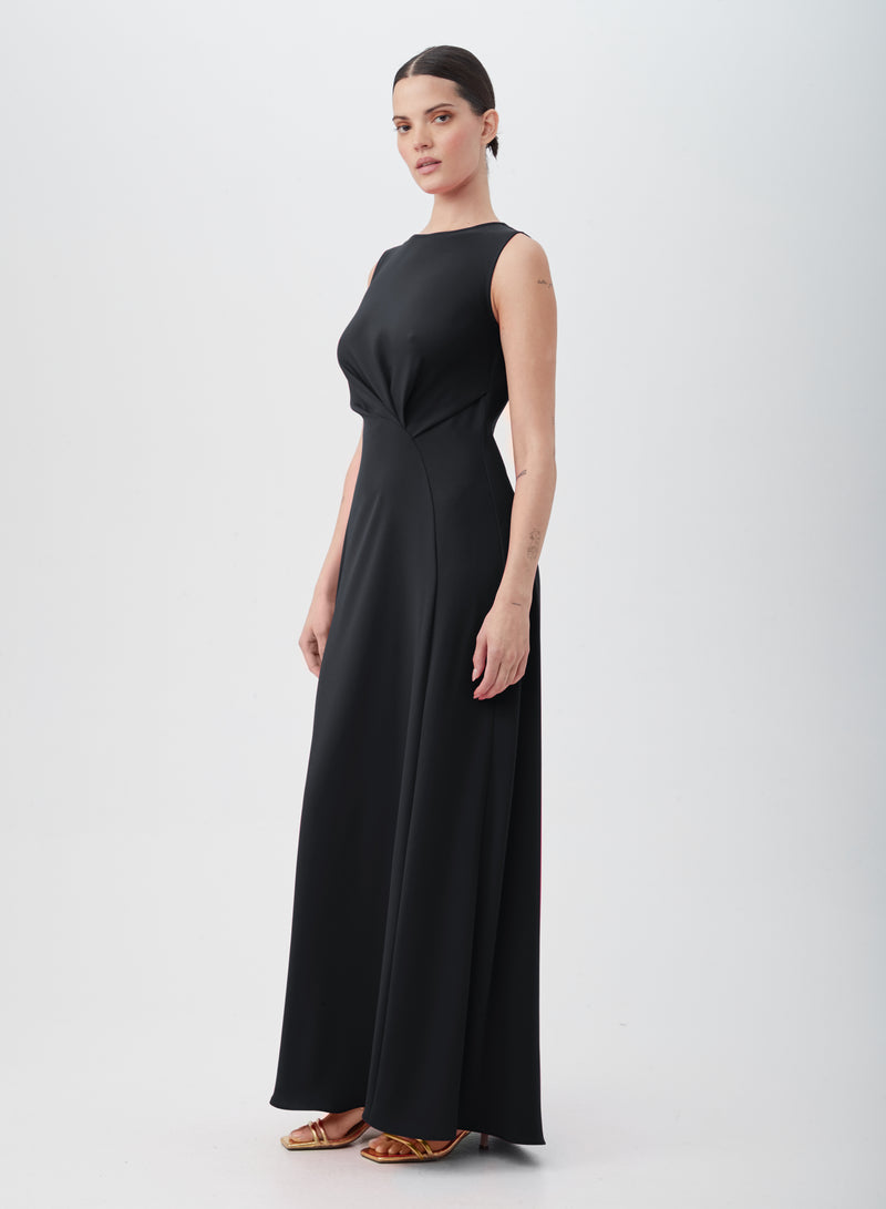 A woman wears the MYTHICAL DRESS, a sleeveless, floor-length black gown of 100% polyester with a fitted waist, paired with gold high-heeled sandals, posed against a plain white background.