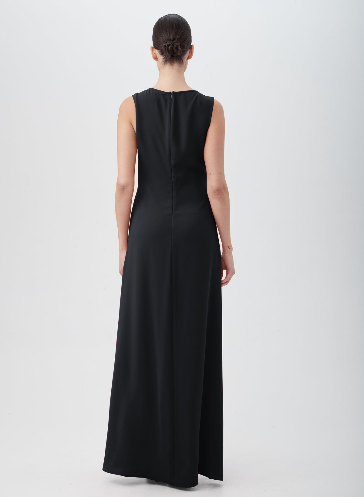 A person with a low bun stands facing away, wearing the floor-length, sleeveless MYTHICAL DRESS in black, made from 100% polyester. The background is plain and light-colored.