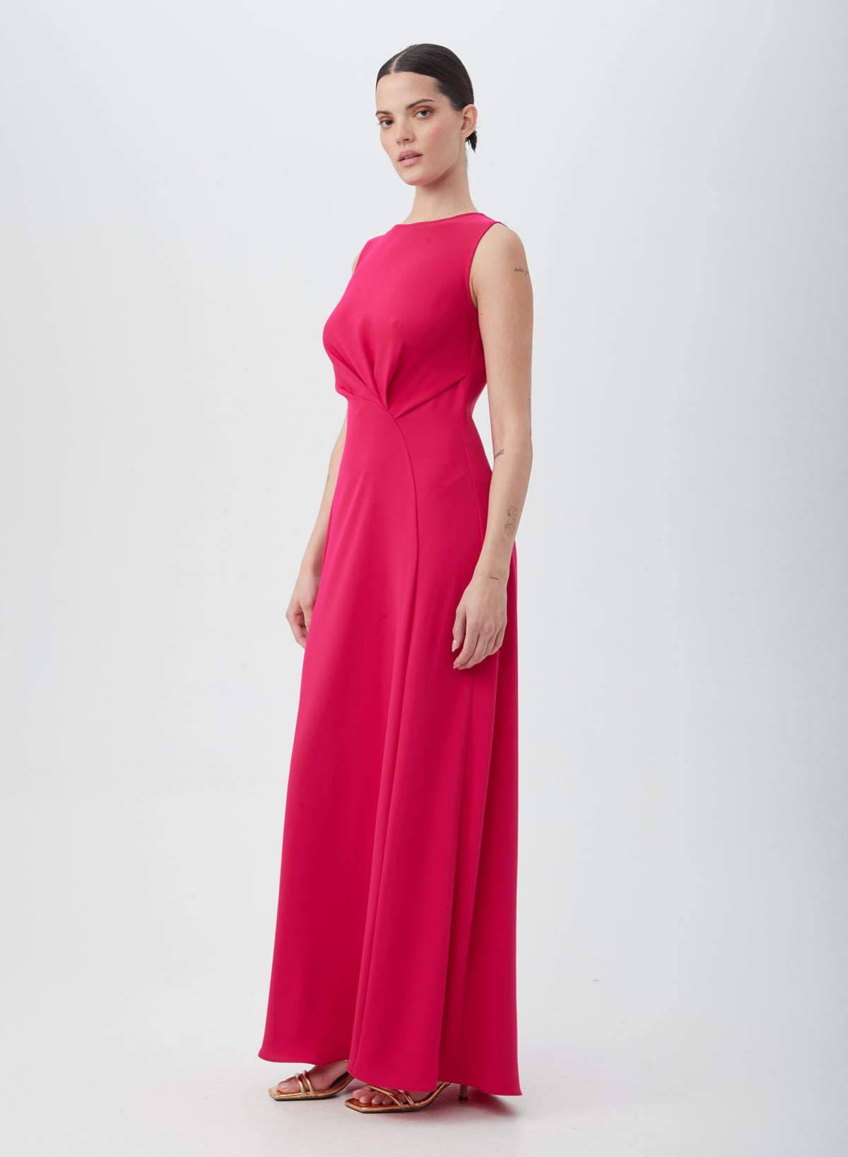 A woman poses against a white background in the MYTHICAL DRESS, a sleeveless, floor-length bright pink dress made of 100% polyester with a fitted bodice and flowing skirt, styled with gold strappy heels.